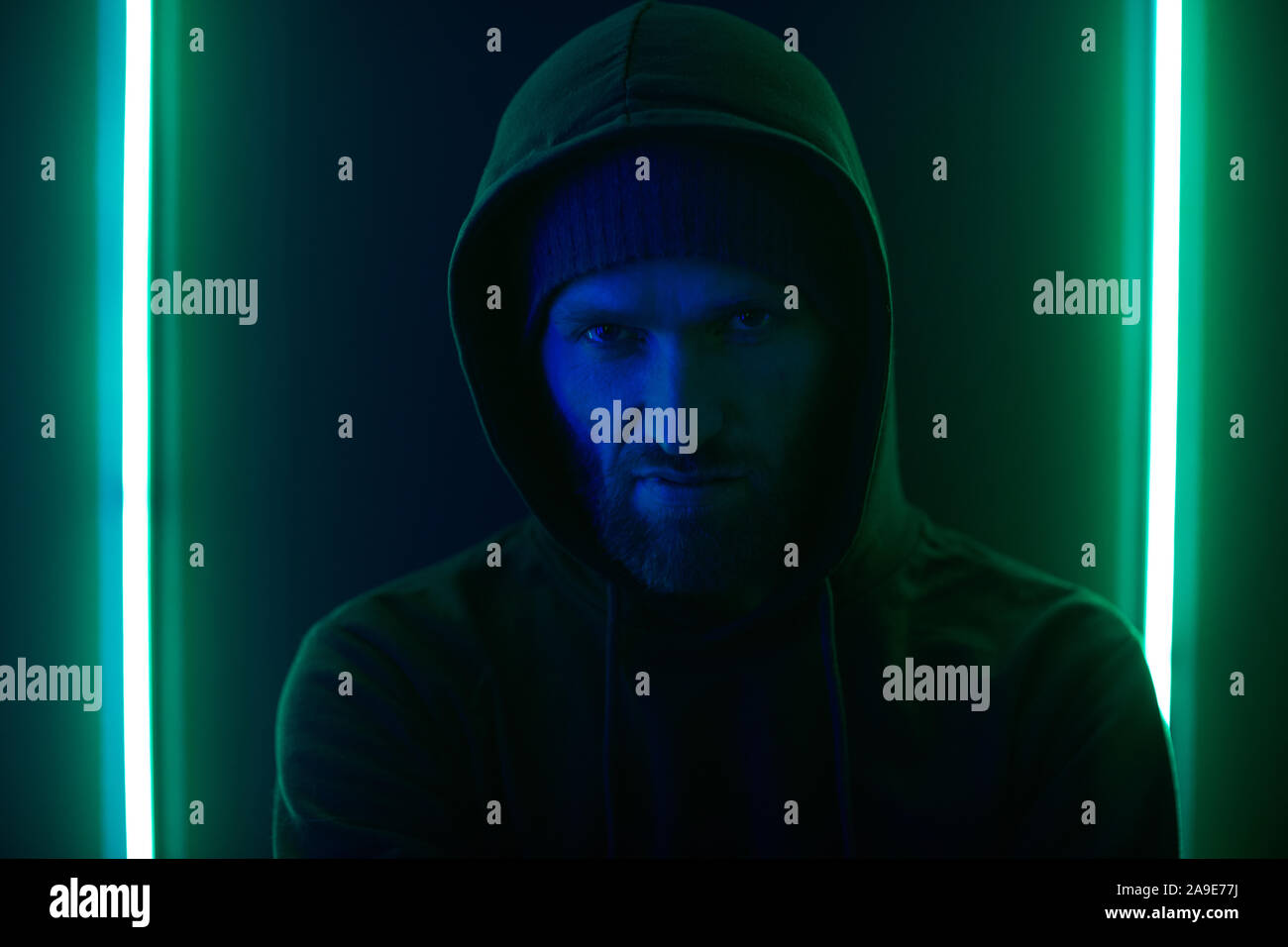 Portrait of young bearded man in dark clothing wearing hoody standing against the wall with green light Stock Photo