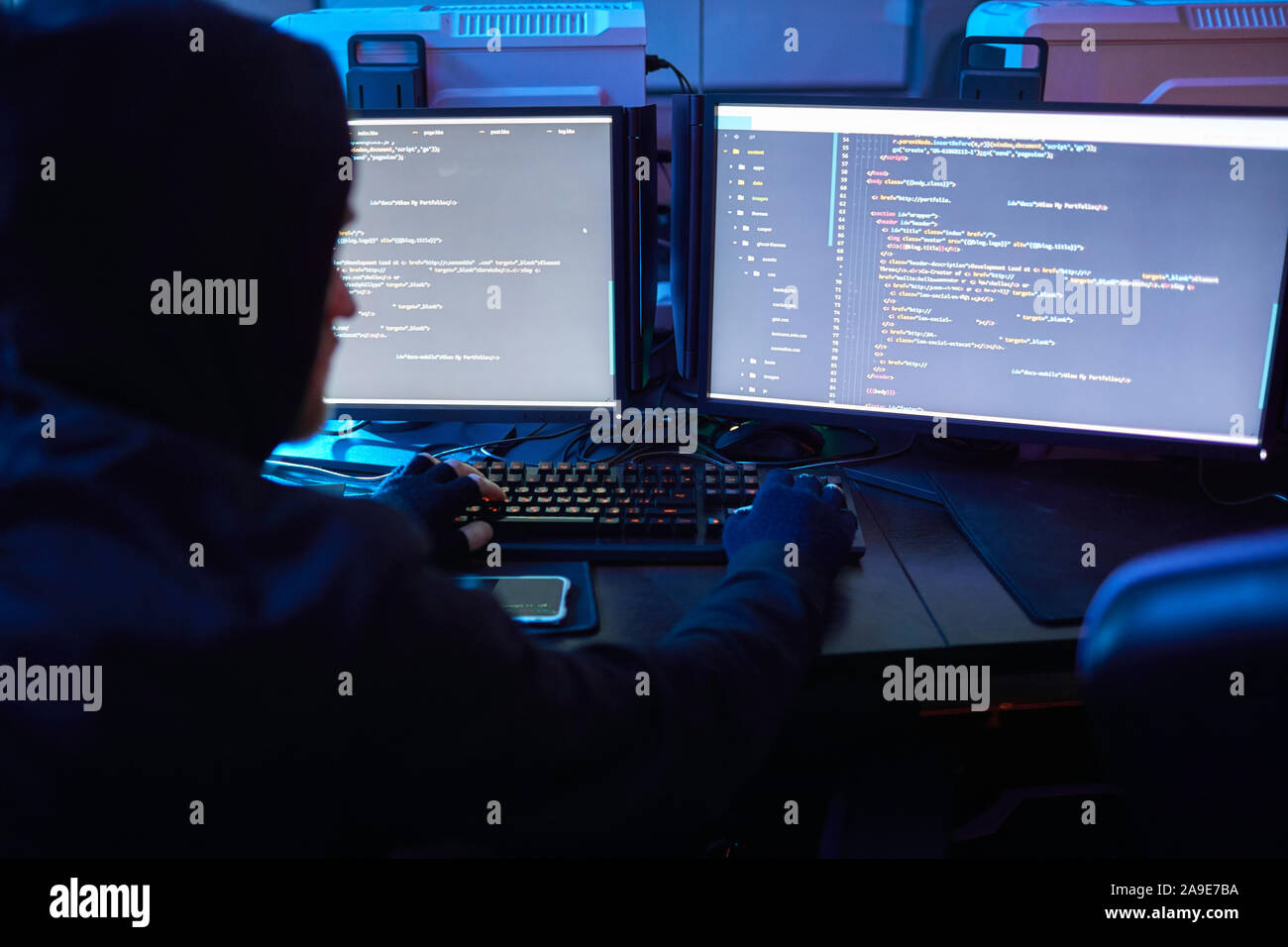 Rear view of computer hacker wearing hoody sitting at the table in front of computer monitors and breaking the computer codes in dark office Stock Photo