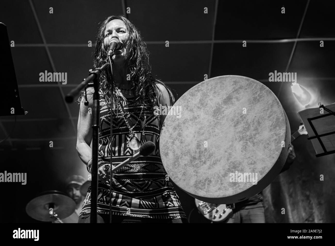 A female musician is viewed from a low angle as she sings and playing native American traditional drum, blurry band members are seen performing in background Stock Photo