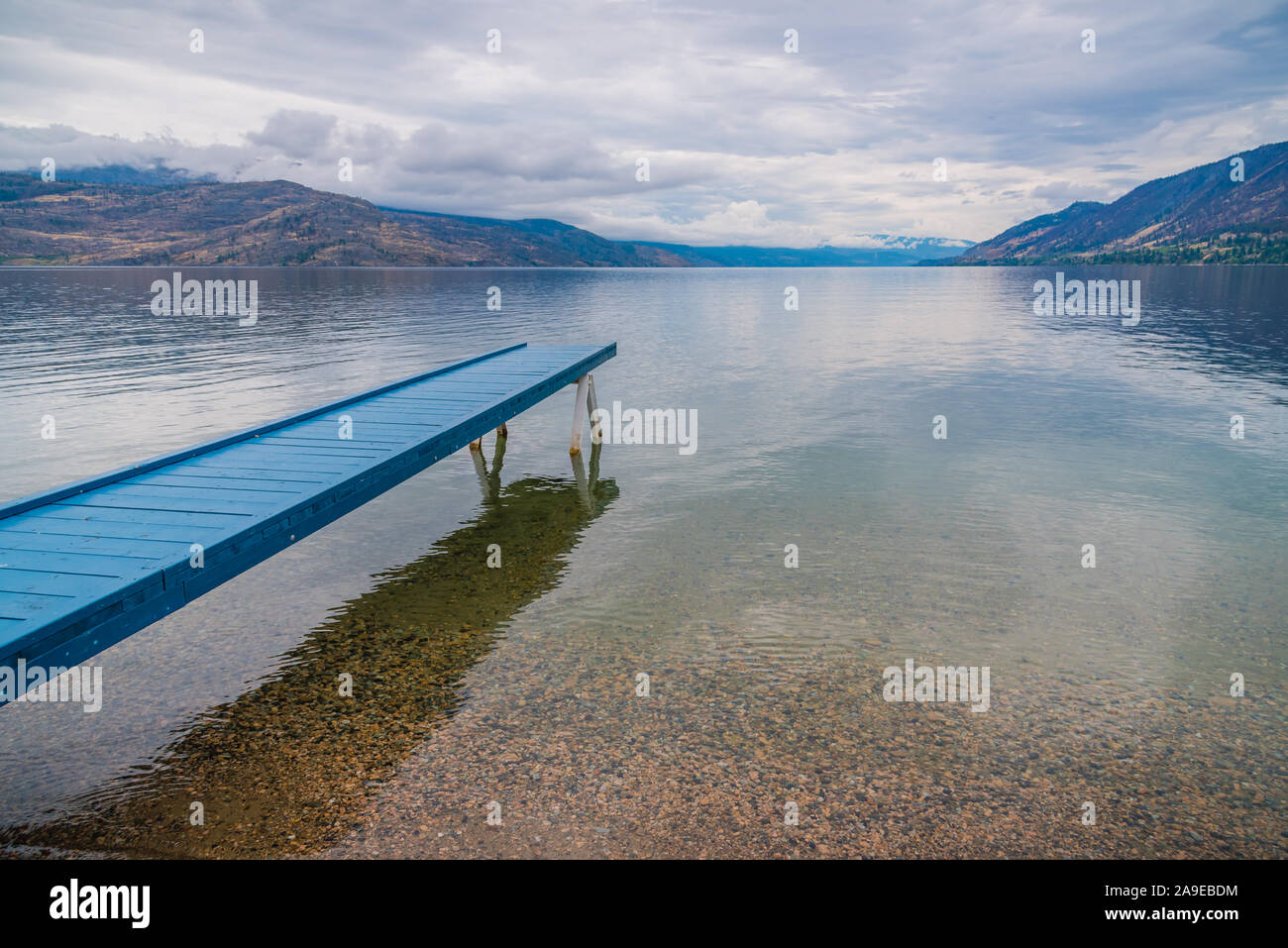 Blue painted dock extending over calm lake with view of overcast sky and mountains Stock Photo