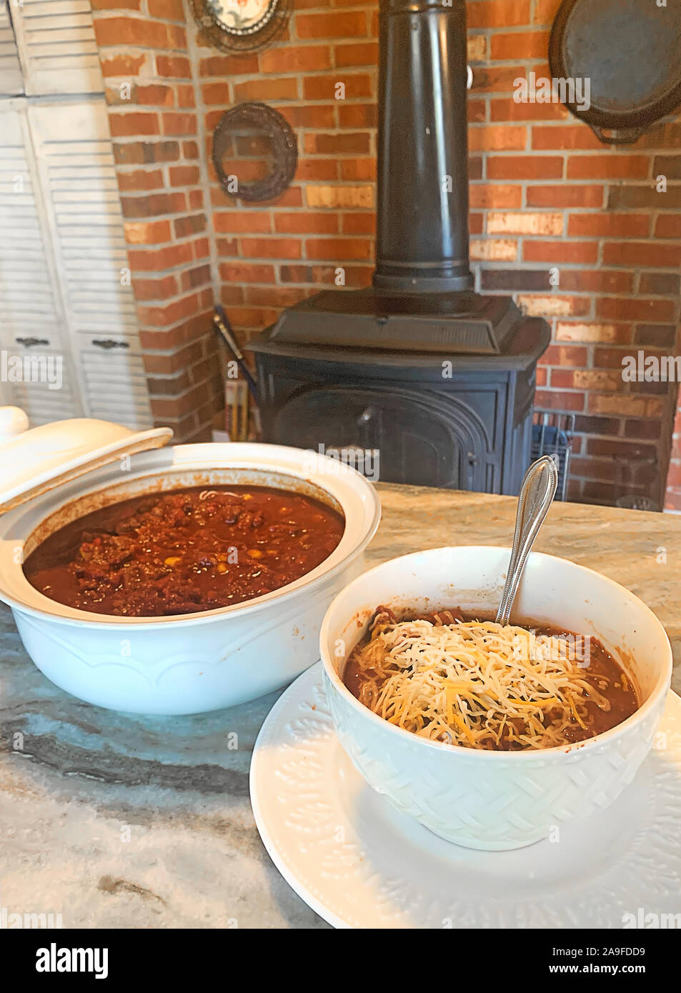 Warm and cozy image of a serving dish filled with chili and also a bowl of Chili with Shredded Cheese.  View of Kitchen's wood burning stove in backgr Stock Photo