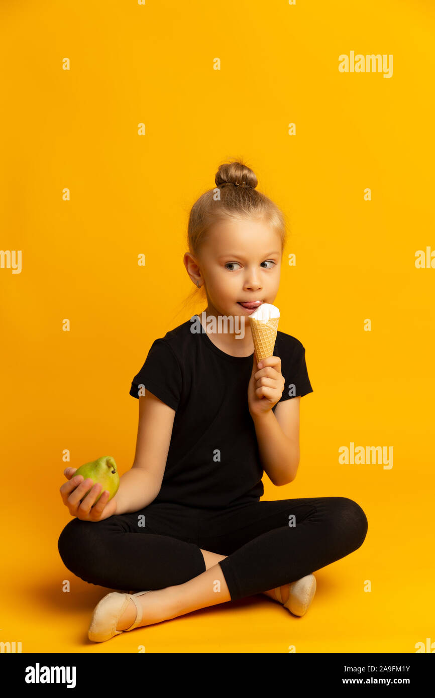 little gymnastic girl in sports clothing holding green pear in one hand and bites an ice cream Stock Photo
