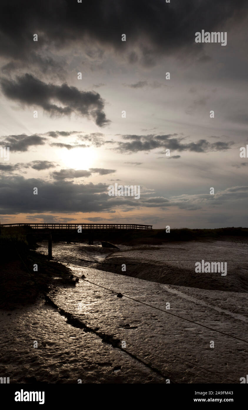 An atmospheric moody salt marsh at low tide with a foot bridge crossing a muddy dyke Stock Photo
