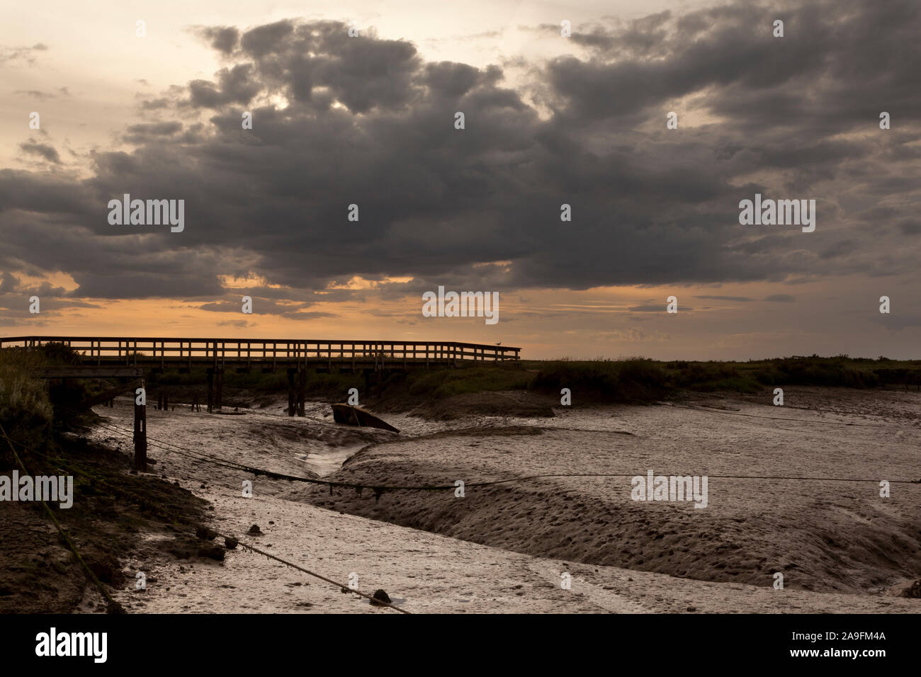 An atmospheric moody salt marsh at low tide with a foot bridge crossing a muddy dyke Stock Photo