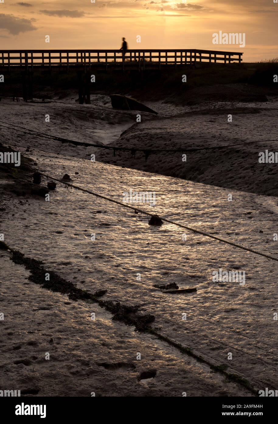 Moody image of Salt Marsh mud at low tide with a shadowy figure walking over a footbridge Stock Photo