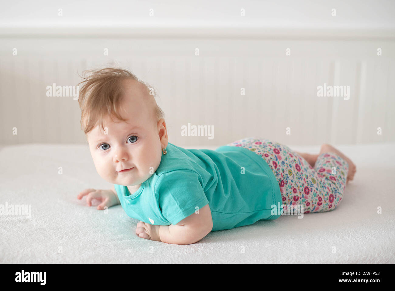 Adorable Caucasian baby girl with blue eyes trying to stand up and walk, looking at the camera calmly with a smile; cute baby facial expressions Stock Photo