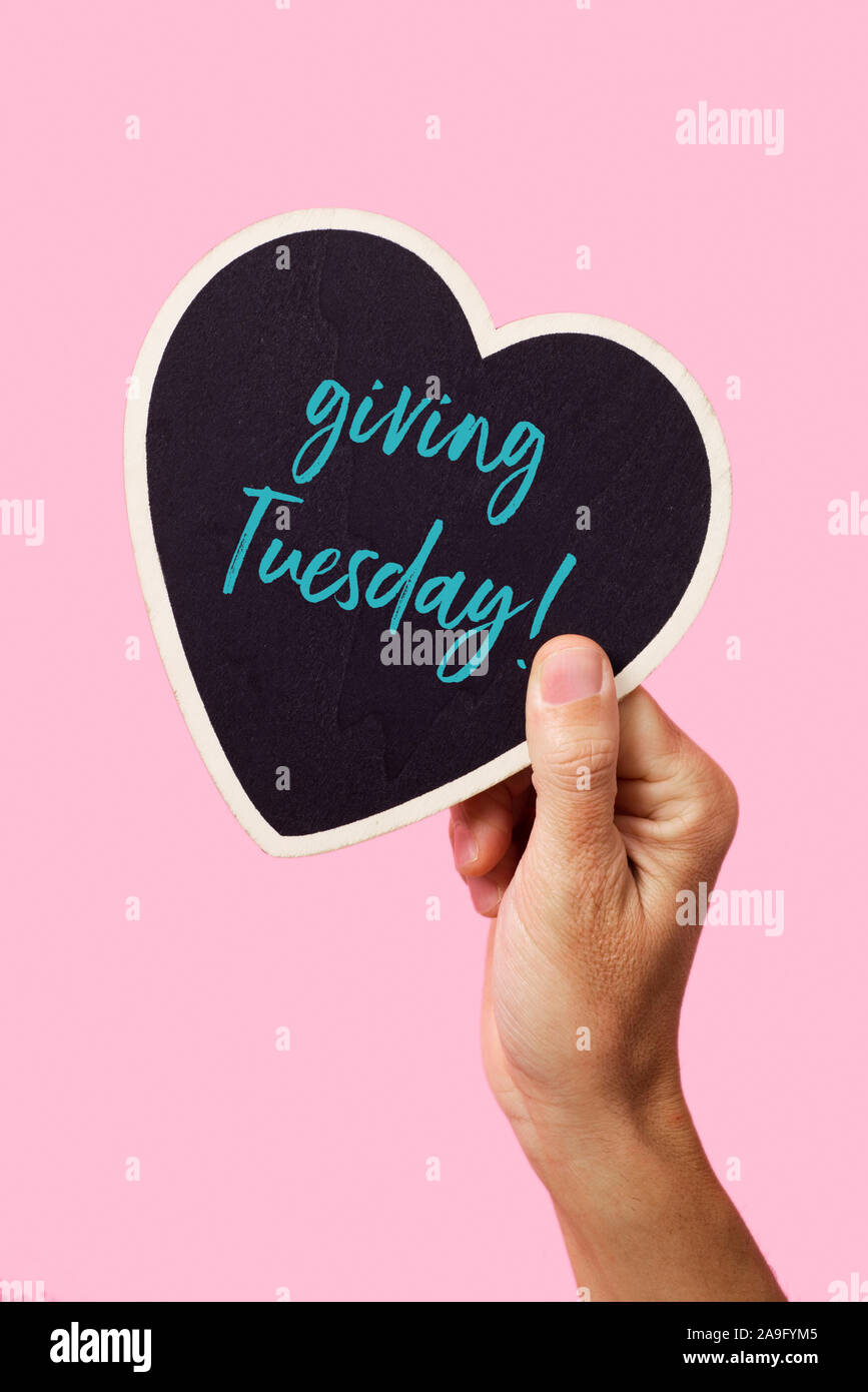 the hand of a young caucasian man holding a black heart-shaped sign, with the text giving tuesday written in it, on a pink background Stock Photo
