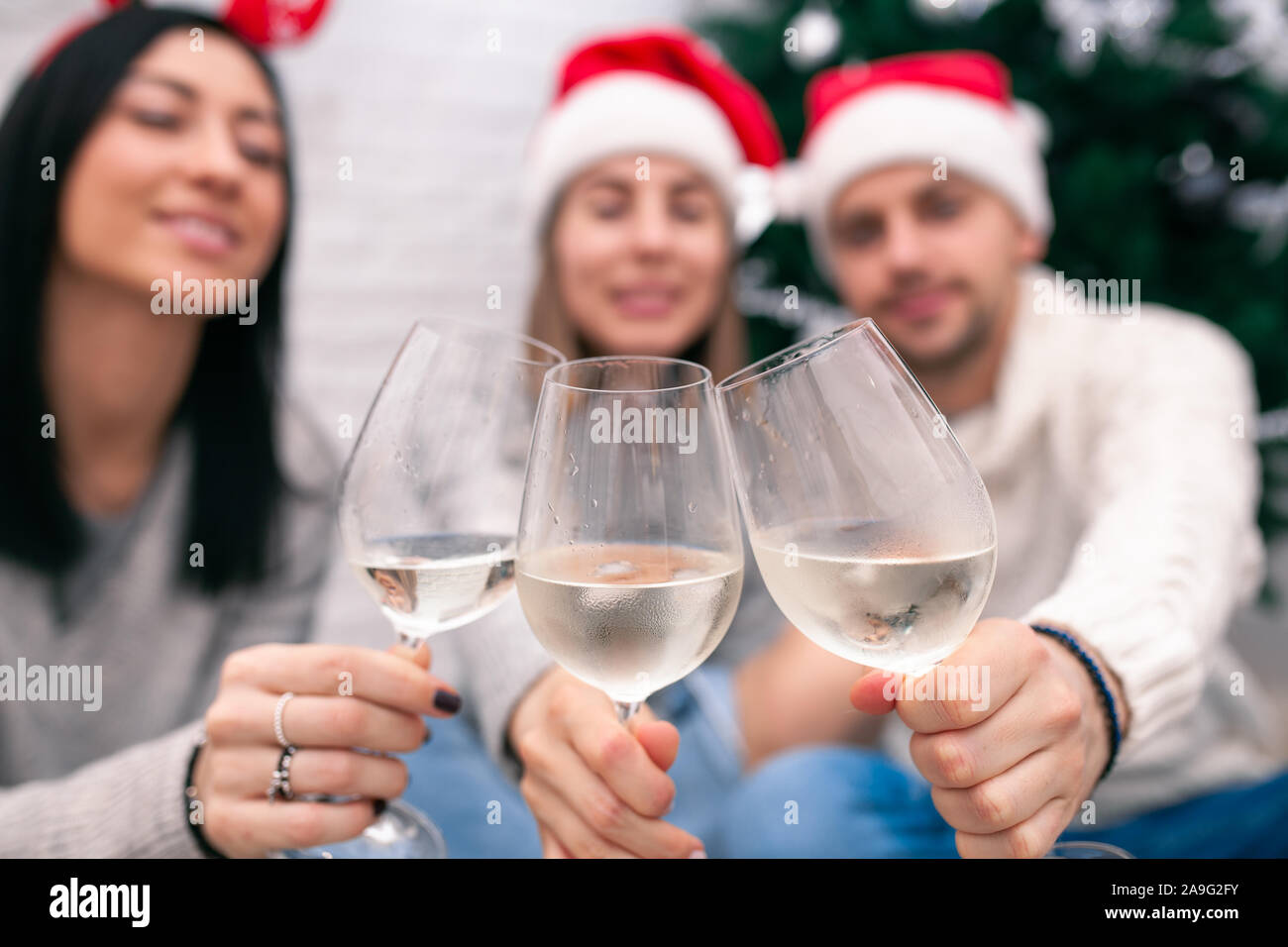 Happy friends celebrating New Year in home interior in Christmas hats sitting near a Christmas tree with glasses of wine. Wine glasses are the focus. Stock Photo