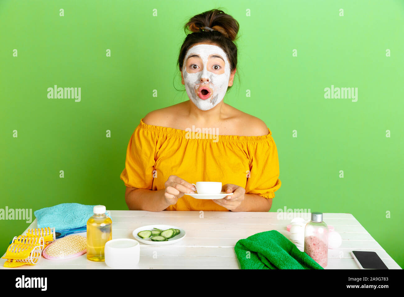 Portrait of young caucasian woman in beauty day, skin and hair care routine. Female model drinking coffee, tea while applying facial mask. Astonished. Selfcare, natural beauty and cosmetics concept. Stock Photo