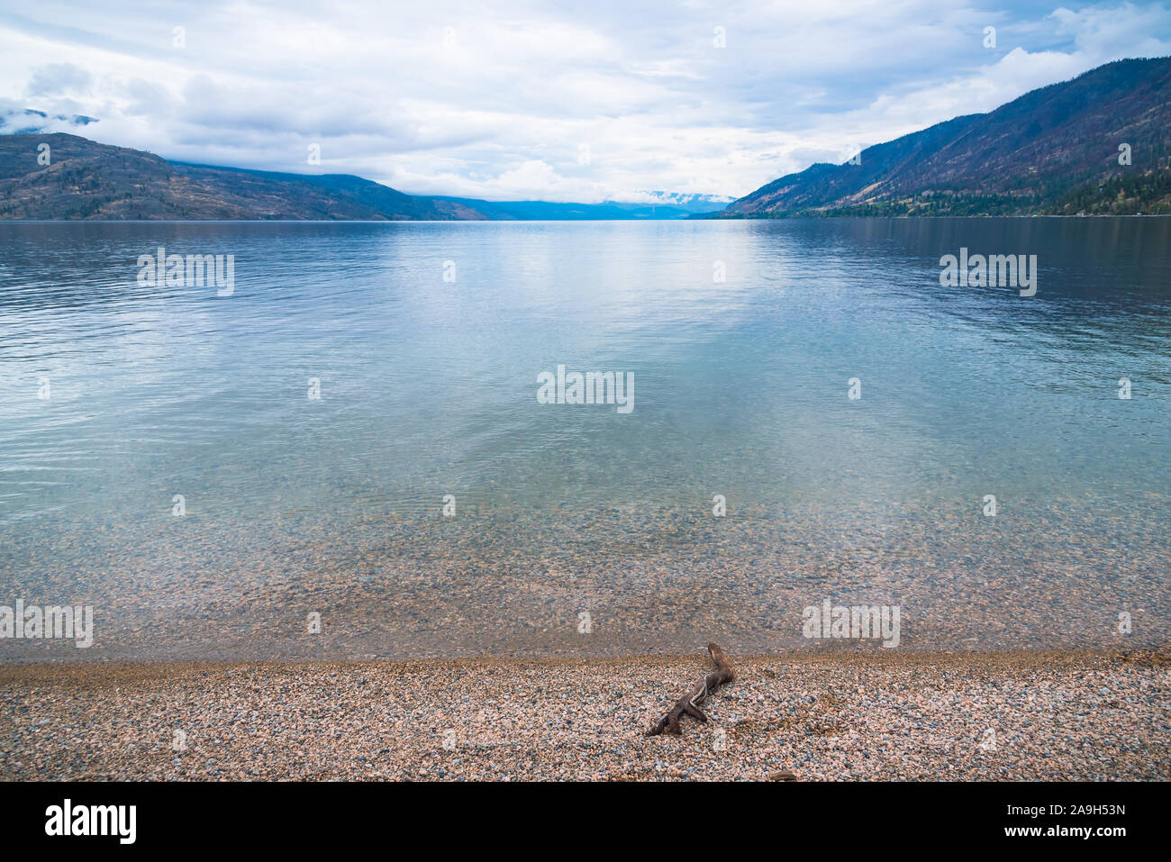 View of pebble beach, calm lake, mountains, and overcast sky at Antler's Beach, Peachland, British Columbia, Canada Stock Photo