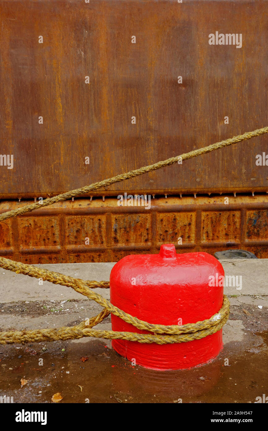 Rope from a ship tied to an old red concrete mooring bollard on a quayside. Stock Photo