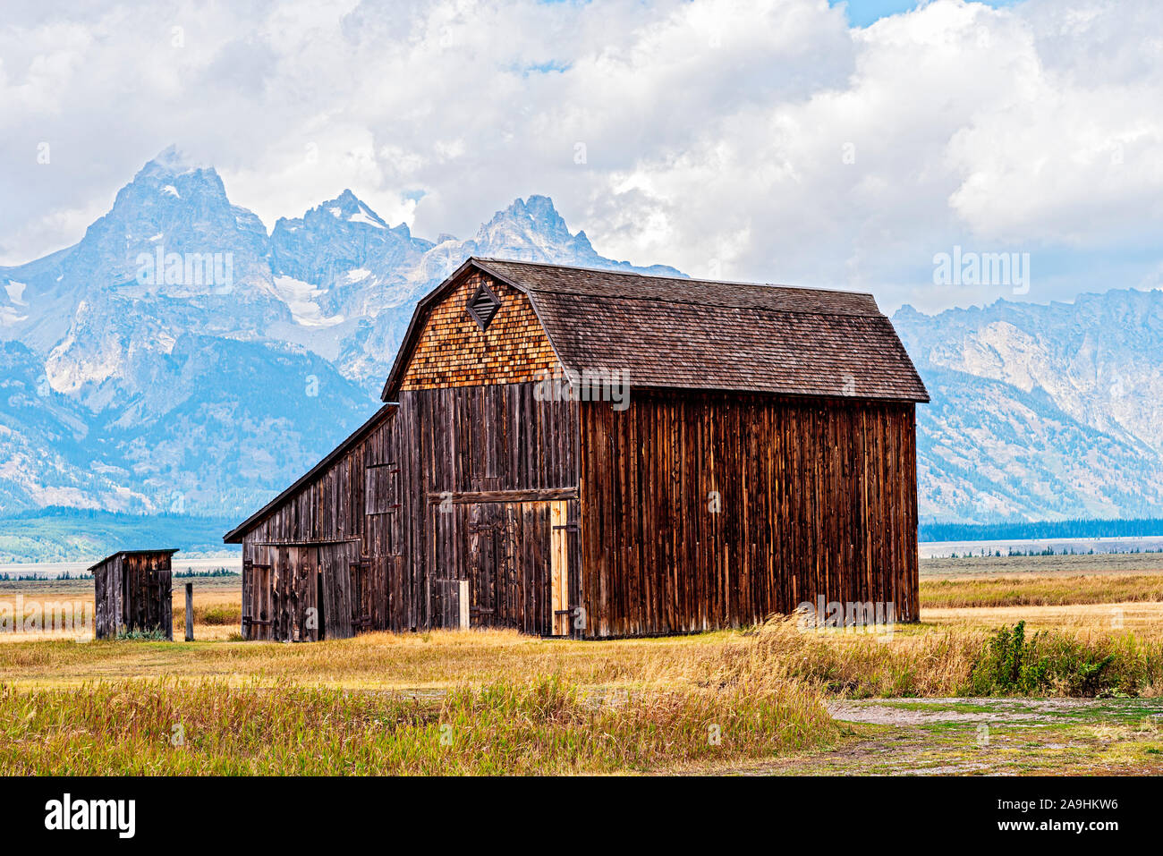 Old abandoned pioneer barn with outhouse behind, grass fields and Rocky Mountains beyond under cloudy skies. Stock Photo