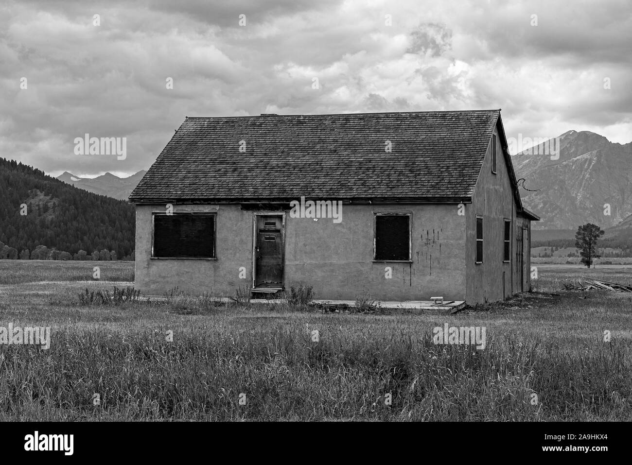 Old abandoned pioneer house in countryside under cloudy skies with mountains beyond, black and white. Stock Photo