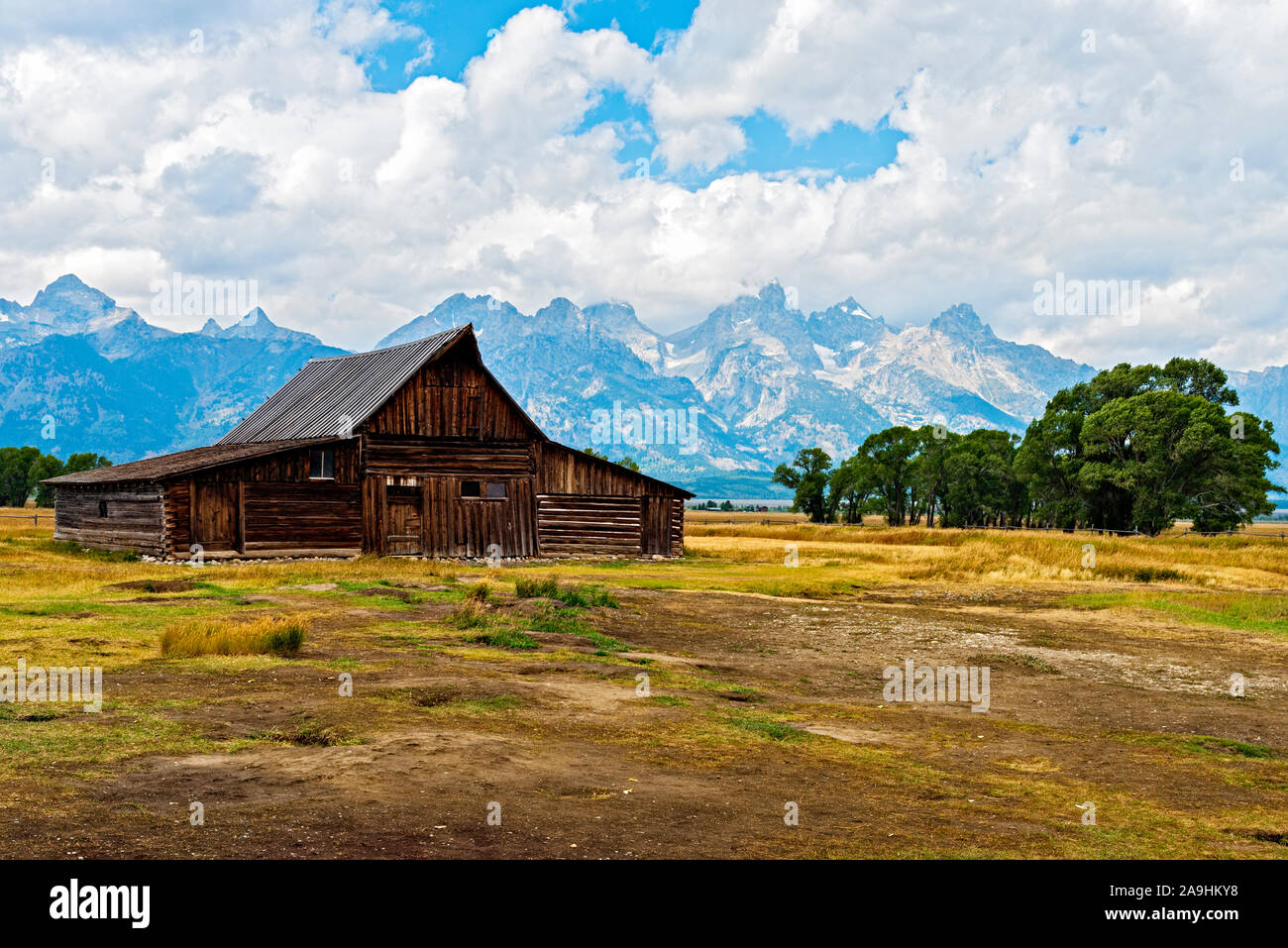 Old Mormon barn, fields, grove of trees and tall Rocky Mountains under a sky with white fluffy clouds. Barn from 1800's. Stock Photo