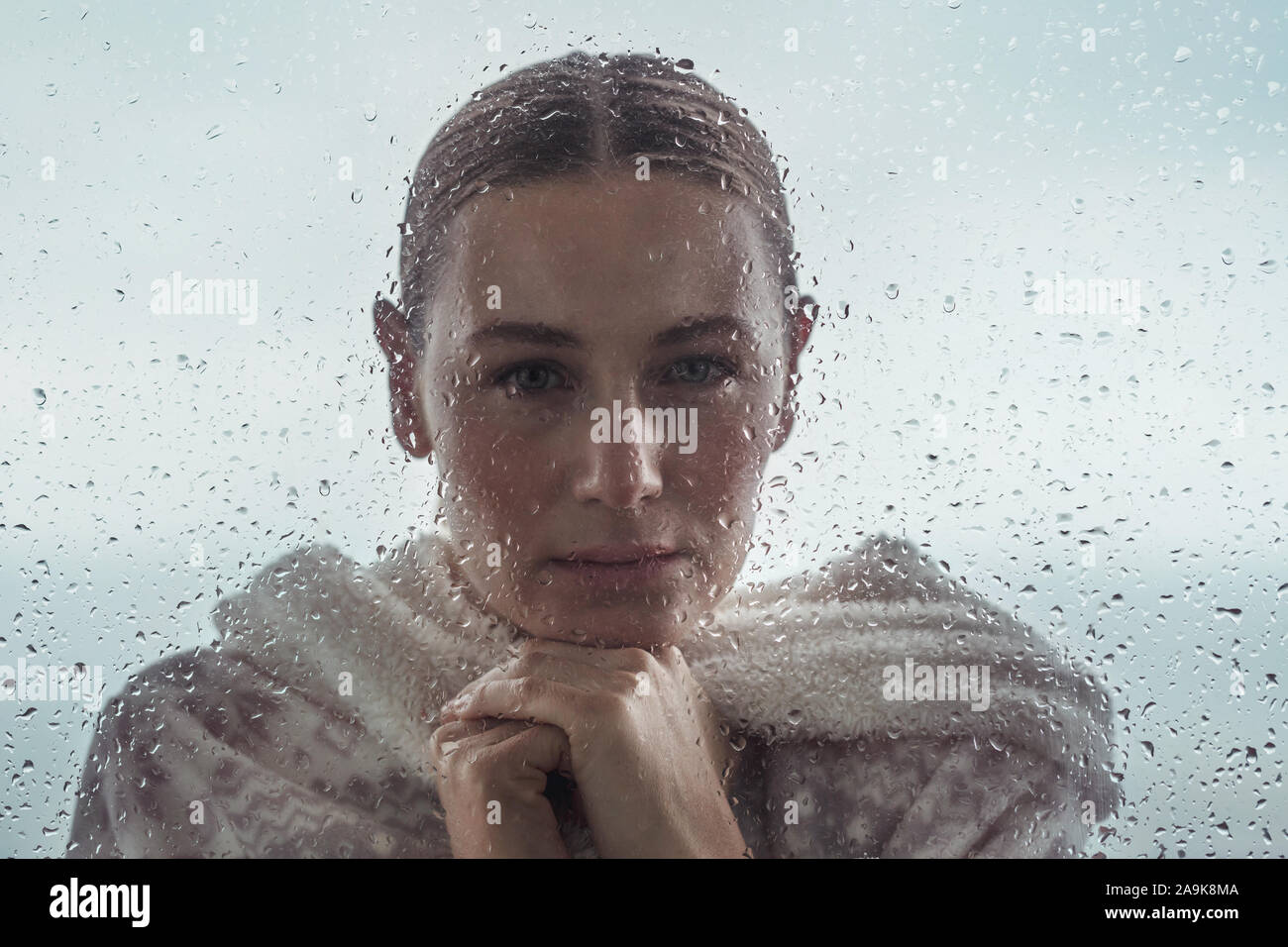 Portrait of a sad woman looking from outdoors into the house through a window covered with raindrops, authentic portrait of a calm girl Stock Photo