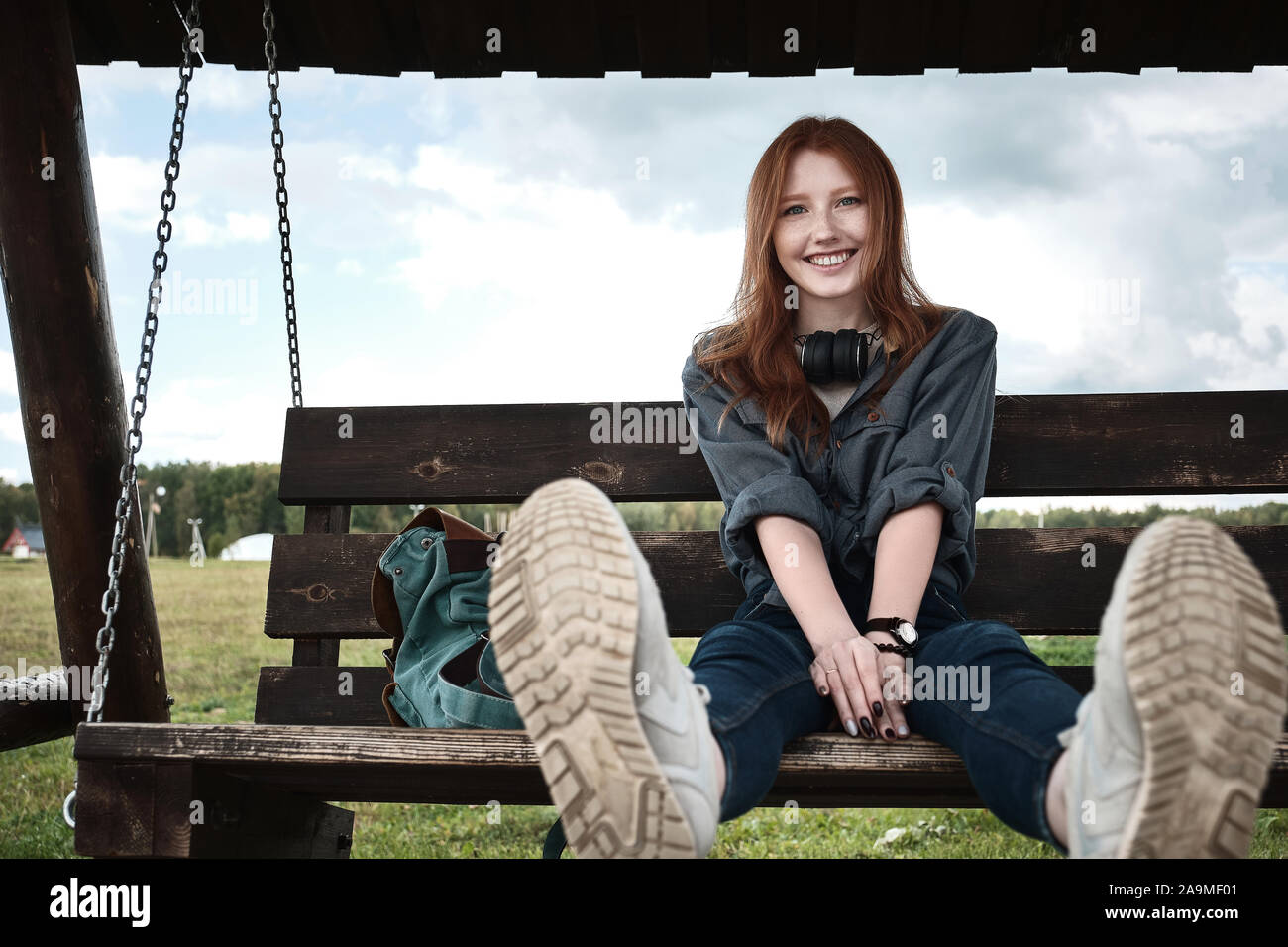 Red-haired girl in a denim shirt sits with a backpack on a wooden swing bench, sways, legs up, smiles broadly into the frame. Stock Photo