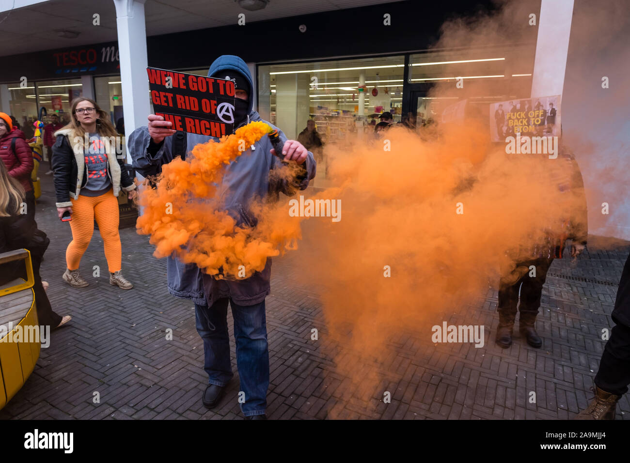 London, UK. 16th November 2019. Anarchists join the protest saying 'Why Vote? Revolt!' and set off an orange flare  as FCKBoris in orange knitted hats walk through Uxbridge shopping centre handing out fliers urging everyone to register and vote against Boris Johnson and kick him out for his racist, elitist politics. They marched with a sound system on a bus to Brunel University to persuade students. Johnson had a majority of just over 5 thousand in 2017 and Labour candidate Ali Milani has strong hopes of beating him and the other 10 candidates. Peter Marshall/Alamy Live News Stock Photo
