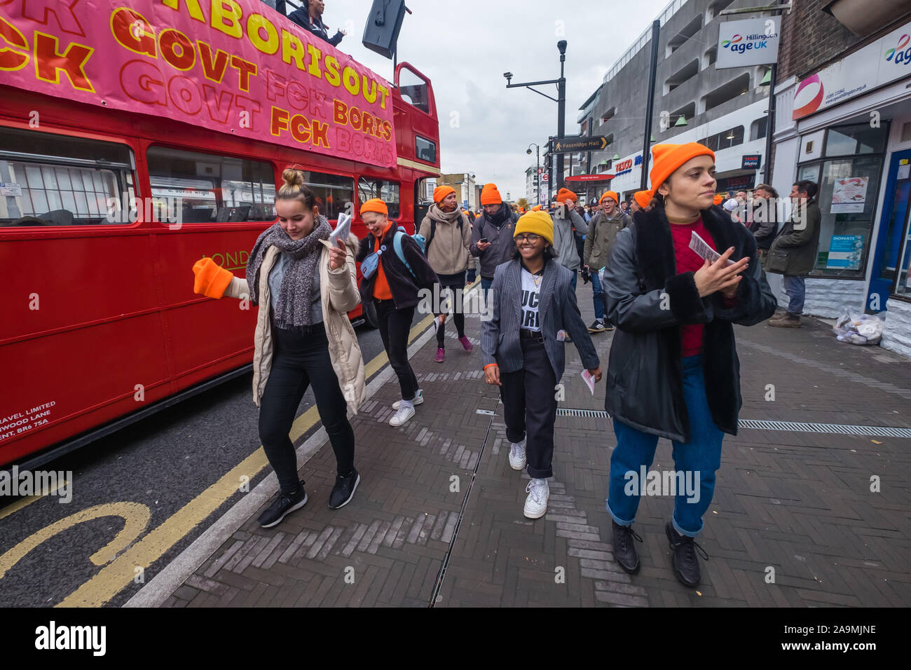 London, UK. 16th November 2019. The bus moves off and people dance along with it. Protesters from FCKBoris in orange knitted hats walk through Uxbridge shopping centre handing out fliers urging everyone to register and vote against Boris Johnson and kick him out for his racist, elitist politics. They marched with a sound system on a bus to Brunel University to persuade students. Johnson had a majority of just over 5 thousand in 2017 and Labour candidate Ali Milani has strong hopes of beating him and the other 10 candidates. Peter Marshall/Alamy Live News Stock Photo