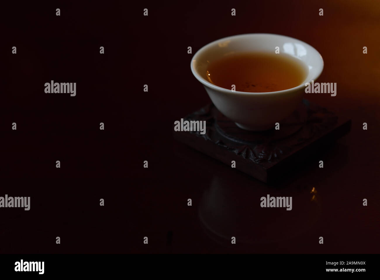 A small white Chinese teacup with black fresh tea in it stands against brown and dark background and is reflected in golden light on a tabletop with s Stock Photo