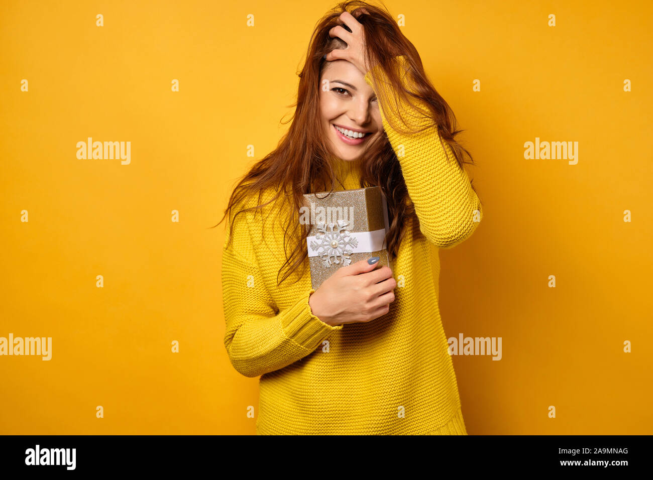 The brunette stands on a yellow background in a yellow sweater, presses a gift box to herself, smiling and ruffling hair with hand Stock Photo