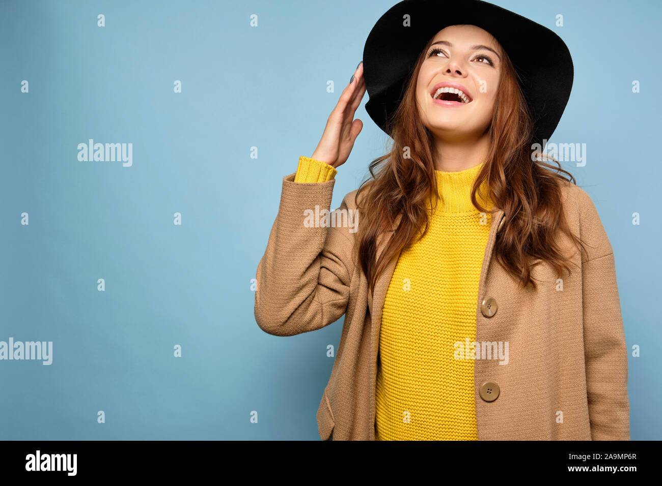 The brunette stands on a blue background in a yellow sweater, trench coat and black hat, joyfully looks at the top. Stock Photo