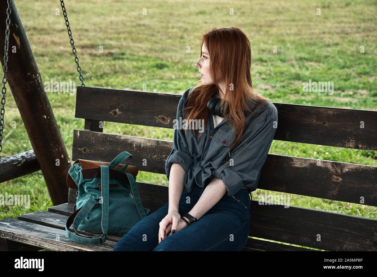 Red-haired girl sits with a backpack on a wooden bench swing and looks away. Stock Photo