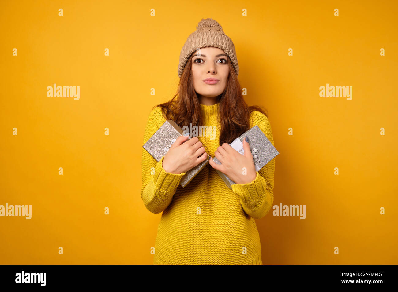 The brunette stands in a yellow sweater and hat, presses gift boxes to herself, looking into the frame with wide eyes. Stock Photo