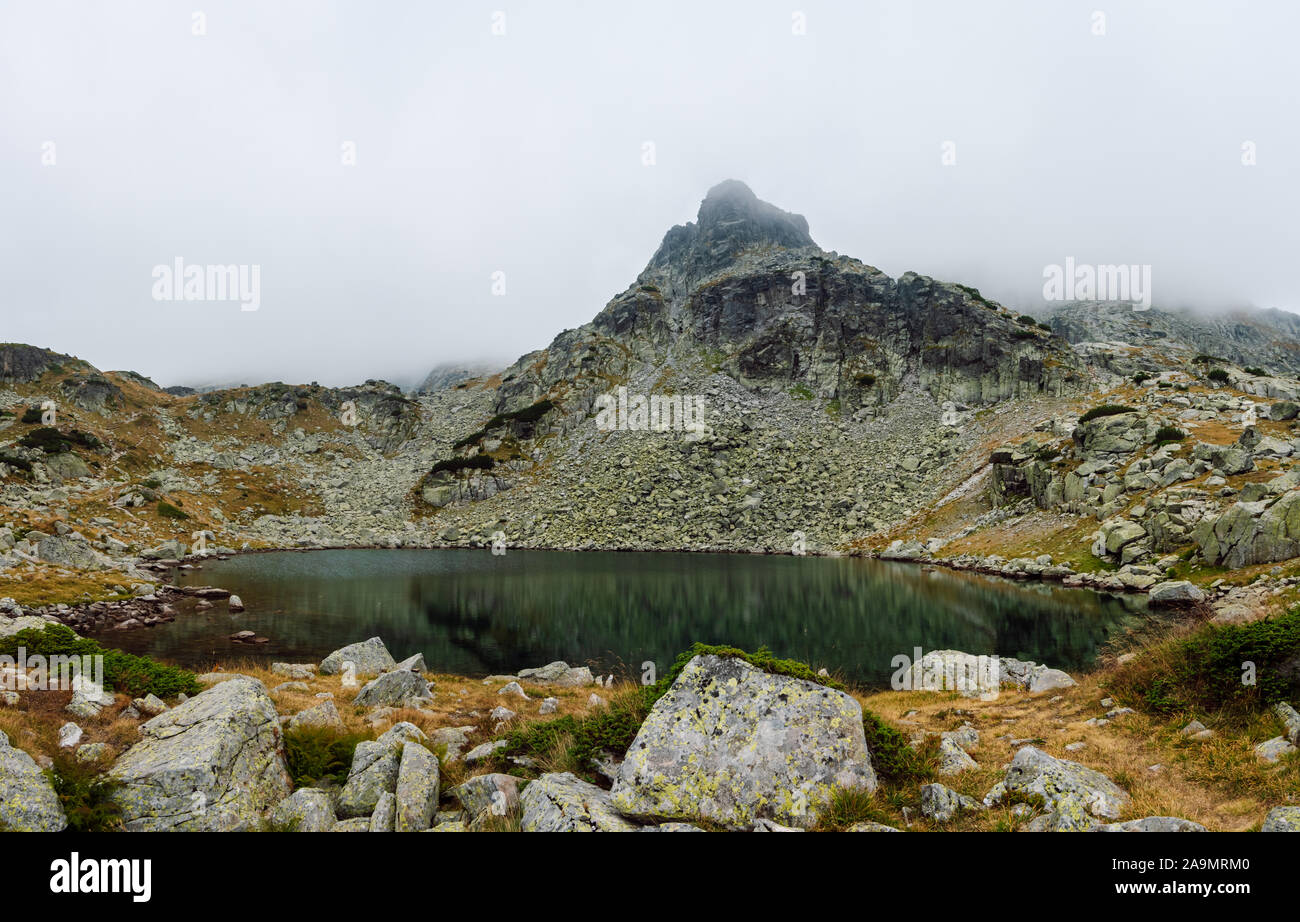 Panoramic view of high mountain lake surrounded by rocks and stone blocks, behind it a rocky peak covered with mists, Rila National Park, Bulgaria. Stock Photo