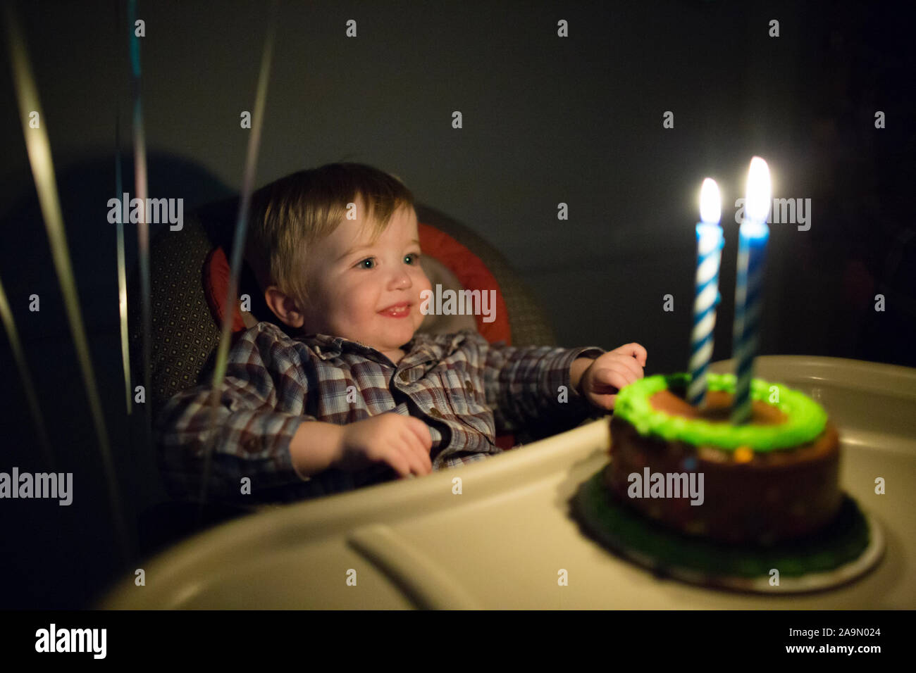 Toddler boy smiles at lit birthday candles on small birthday cake Stock Photo