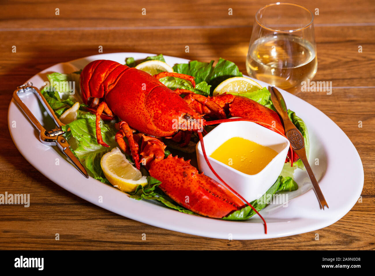 Cooked lobster on a white ceramic platter, romaine lettuce, lemon slices, melted butter, and white wine on the side in a stemless wine glass Stock Photo