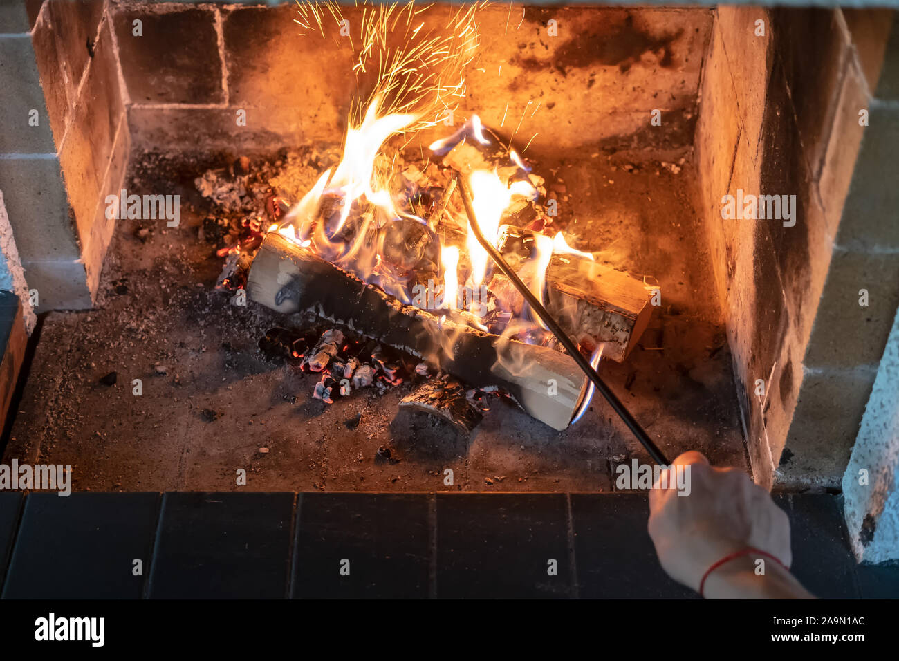 Female hand straightens and overturns burning firewood with an iron poker in an open fireplace and from the coal fly beautiful spark. Warmth and Stock Photo