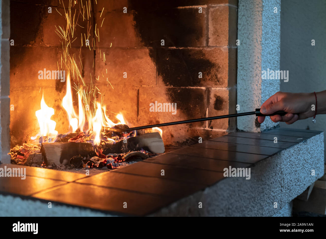Female hand straightens burning firewood with an iron poker in an open fireplace and from the coal fly beautiful spark. Warmth and comfort in a cozy Stock Photo
