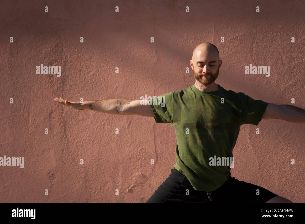 Young man in his thirties with a beard wearing a green tee shirt practicing yoga by a salmon pink colored adobe wall. Stock Photo