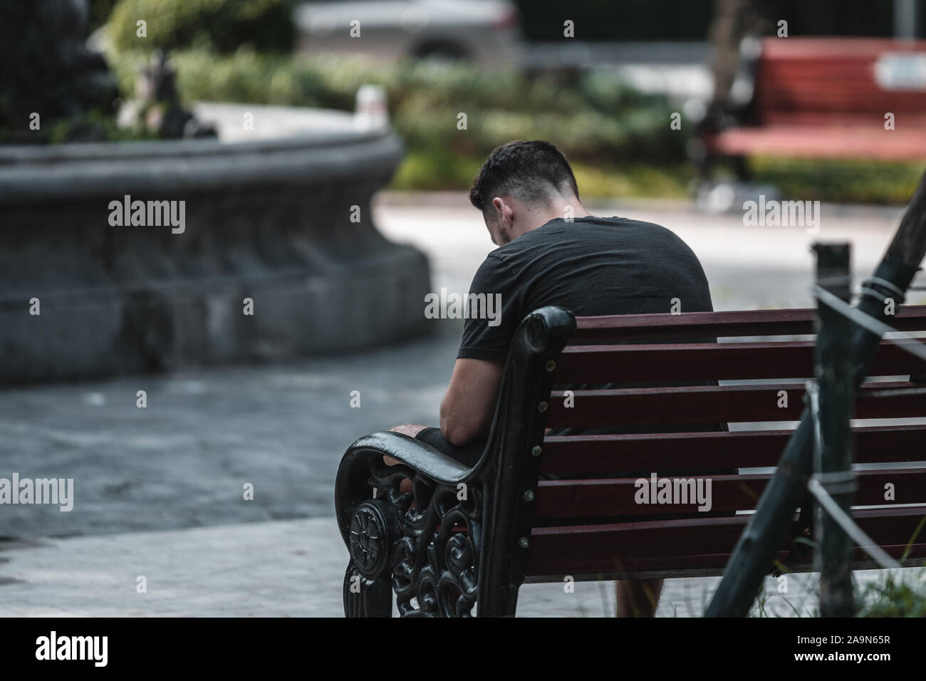 A young white man sits alone on a bench looking down feeling sad Stock Photo