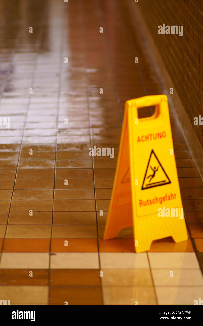 The wet tiles and clinker of a freshly wiped tunnel floor with a warning sign: Caution Slipping hazard! Stock Photo