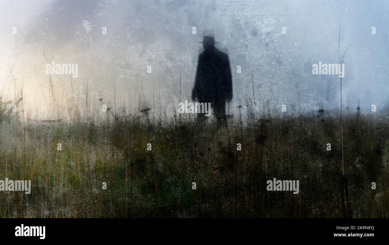 A mysterious figure wearing a long coat and fedora hat, with back to camera, standing in a field. With an artistic, blurred, weathered, textured edit. Stock Photo