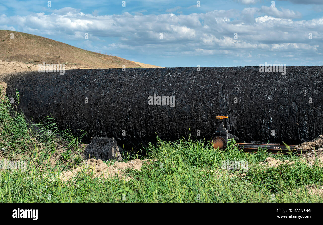 Black Oil pipeline goes out above the ground. Oil-stained pipe. Concept for oil petrol transportation with tubes. Green grass and black color oil pipe Stock Photo