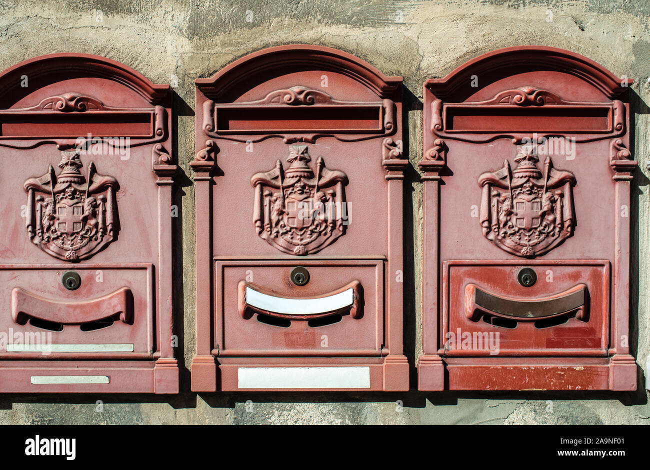 Old vintage mailboxes in Italy. Metal red mailboxes on grey facade. Hard sunlight. Empty plates. Italian house wall. Vintage mail correspondence conce Stock Photo