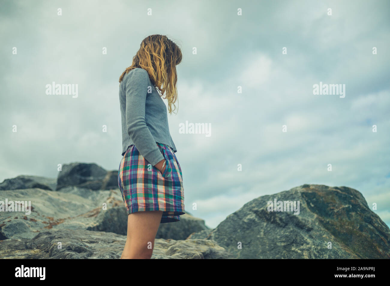 A young woman with her hands in her pockets are standing on some rocks against a cloudy sky Stock Photo