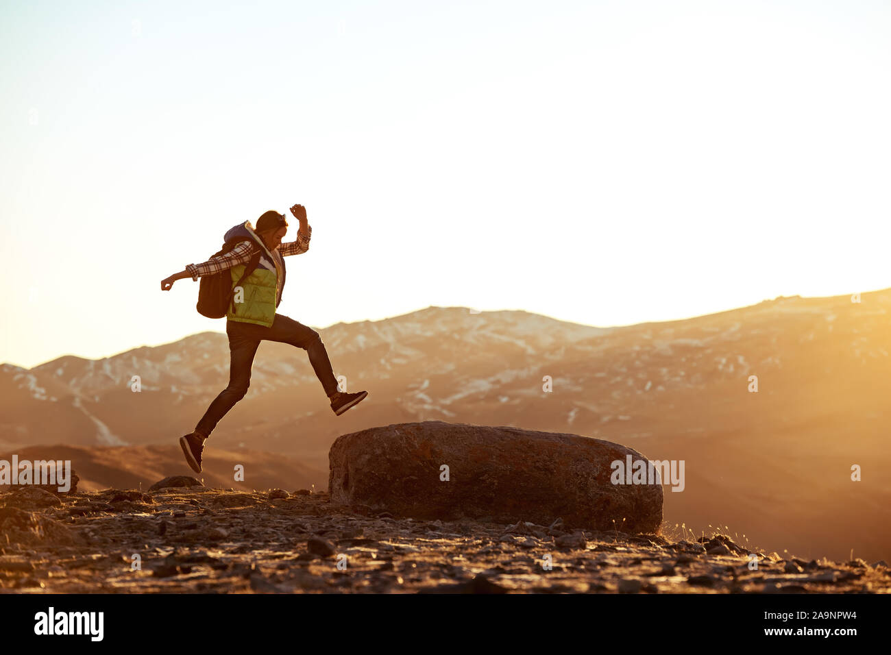 Young sporty girl jumps to big rock against sunset and mountains Stock Photo