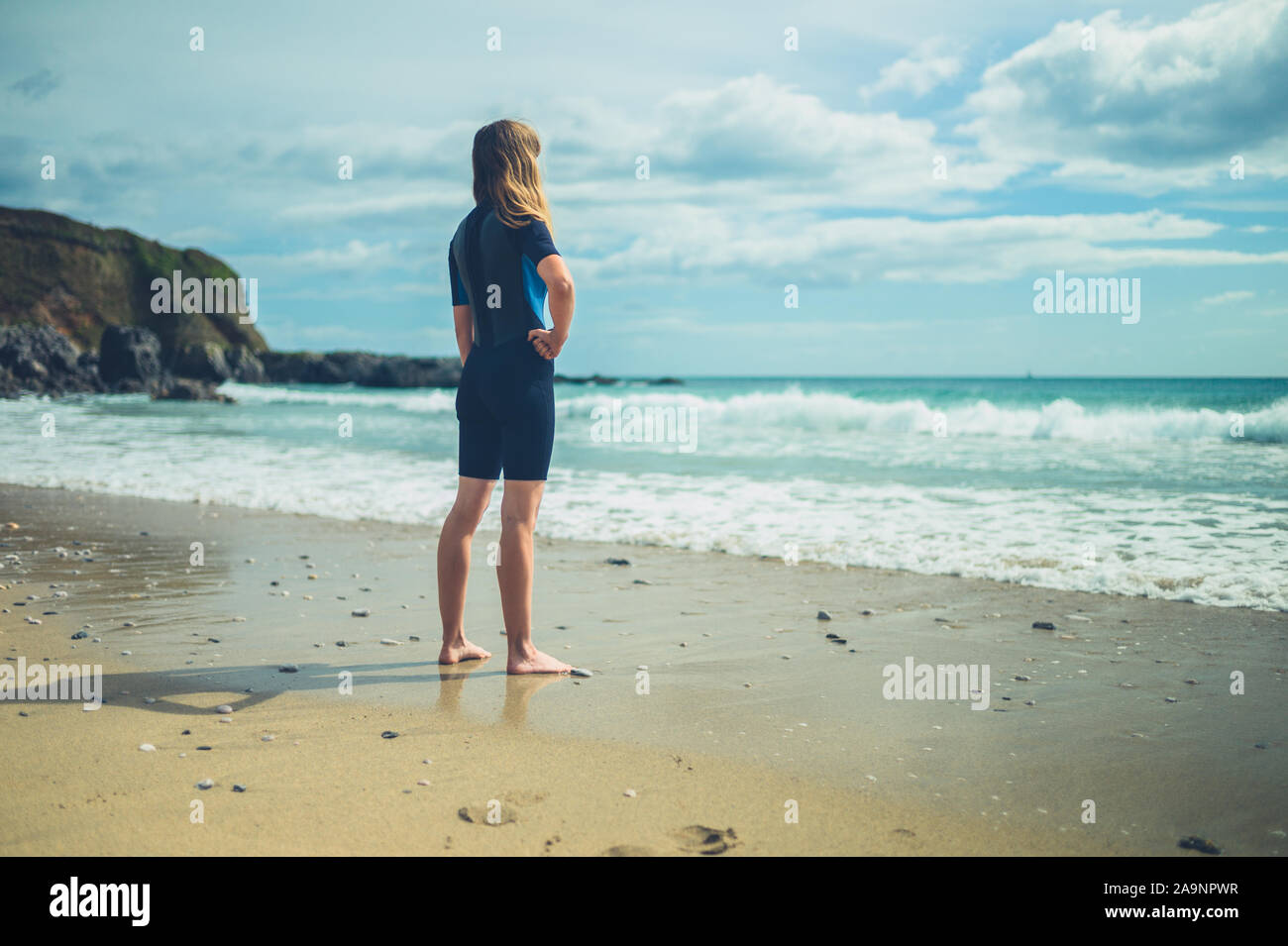 A young woman wearing a wetsuit is standing on the beach in summer ...