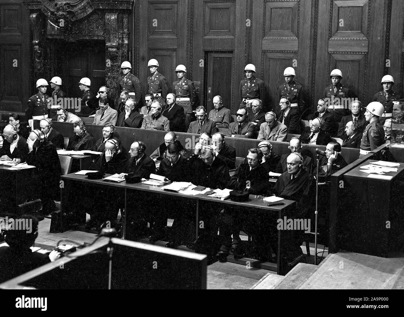 The prosecution charges the defendants with conspiring to destroy the independence of other nations. Goering is in the defendant's box. The defendants, surrounded by American military police, Goering, Hess, Von Ribbentrop, Keiter, Rosenberg, Frank, Frick, Streicher, Funk, Schacht; back row, Donitz, Raeder, Von Schirach, Sauckel, Jodl, Von Papen, Seyss-Inquart, Speer, Von Neurath, and Hans Fritsche. 11/23/45. Stock Photo
