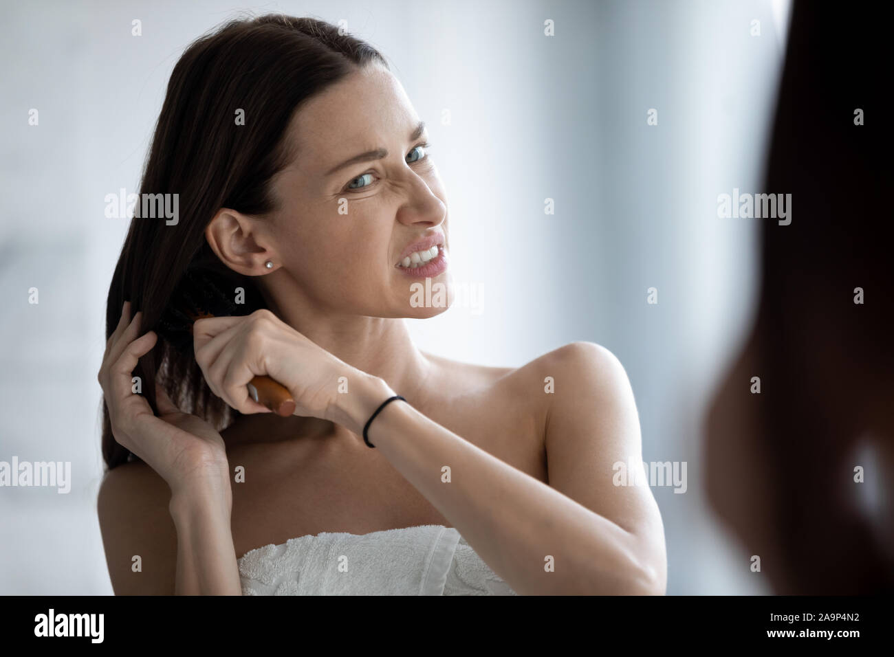 Stressed young lady brushing long tangled hair looking in mirror Stock Photo