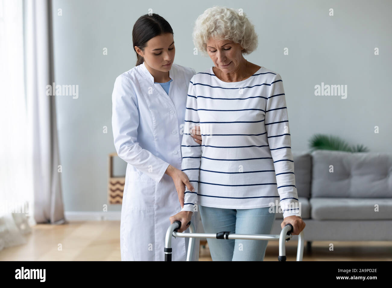 Young woman caregiver helping disabled old grandma patient using walker Stock Photo