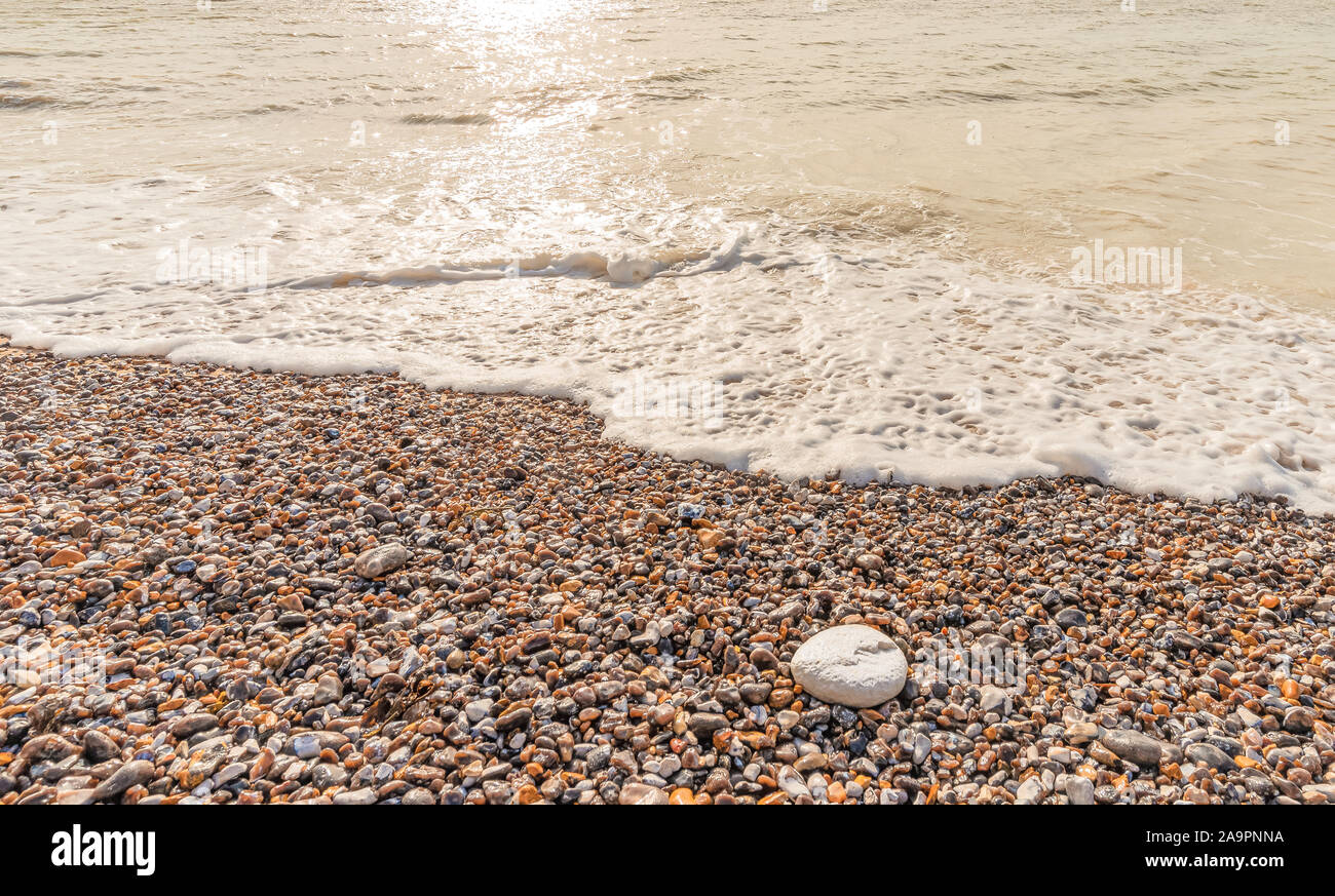 The sea lapping against a beach with multi-coloured pebbles. A low sun is reflected on the surface of the sea. Stock Photo