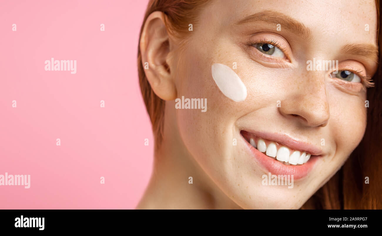 Cropped close up portrait of young woman with cream on cheek. Charming caucasian girl with red hair, fresh freckled skin, smiling happily standing aga Stock Photo