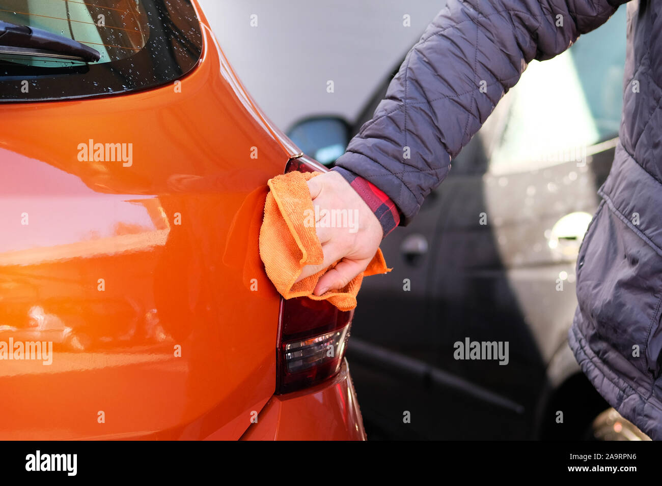 Man after cleaning wipes his orange car with a rag  at car wash. Male hand and car body closeup. Stock Photo