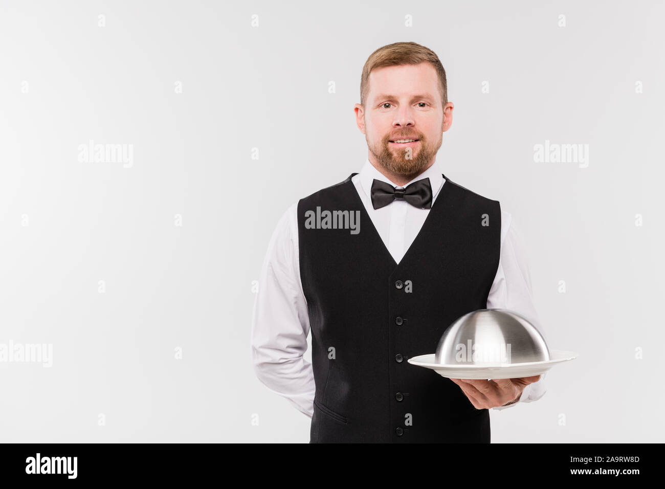 Happy young waiter in waistcoat and bowtie holding cloche with meal Stock Photo