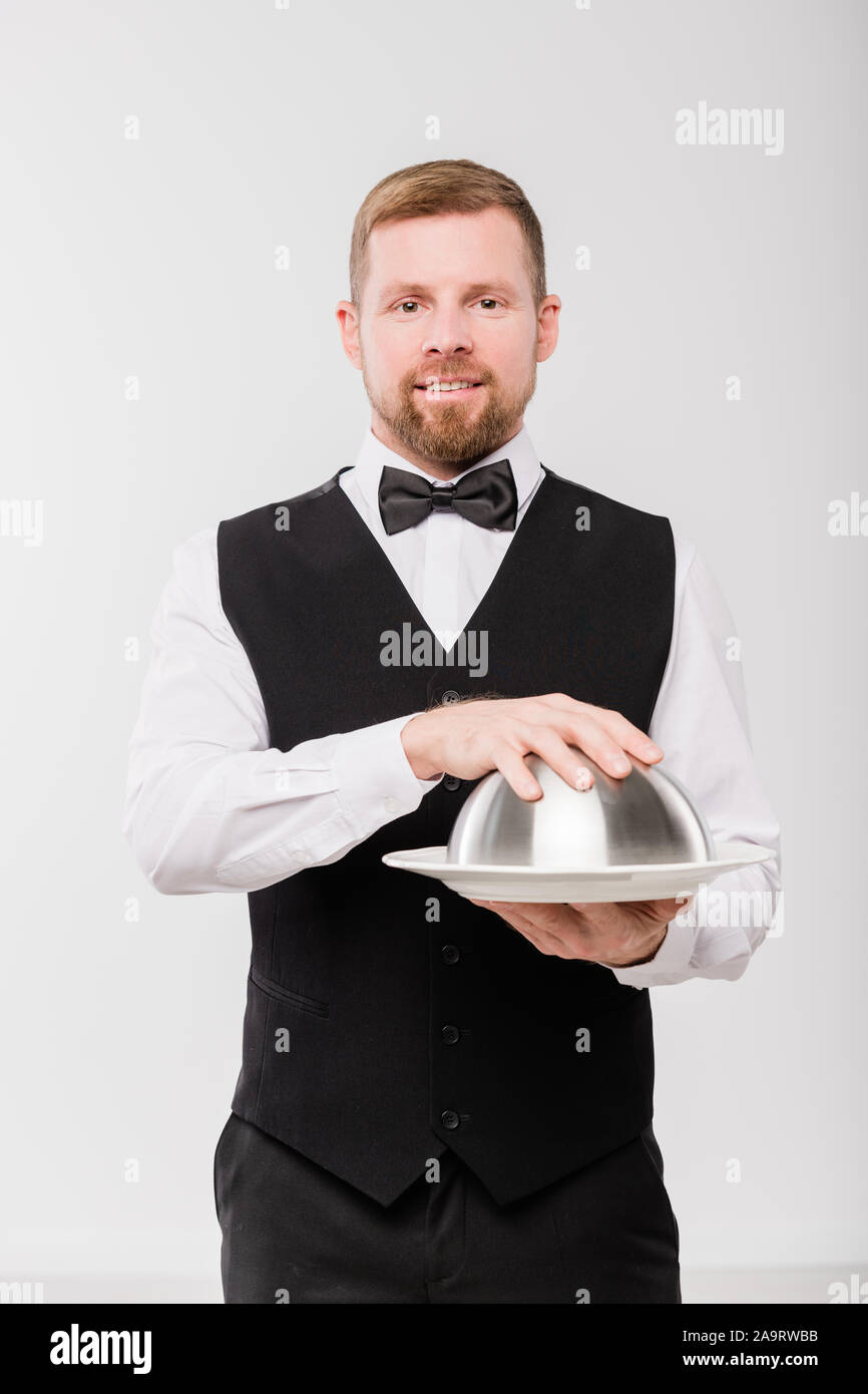 Young elegant waiter in black waistcoat and bowtie holding cloche with meal Stock Photo