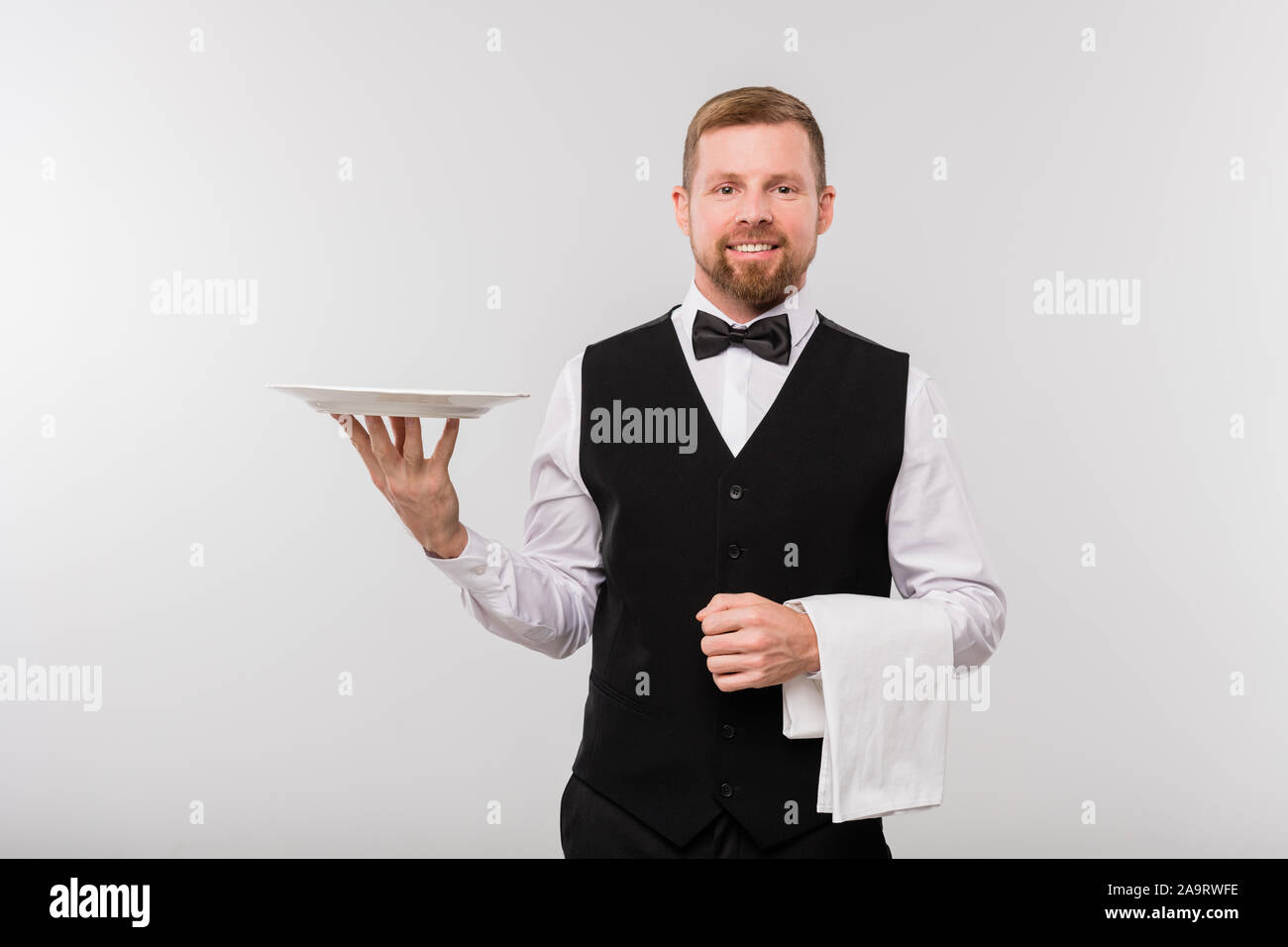 Young elegant waiter in black waistcoat and bowtie holding white towel and plate Stock Photo
